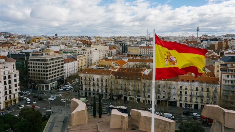 Drone view of Spanish city with aged buildings and national flag under cloudy blue sky