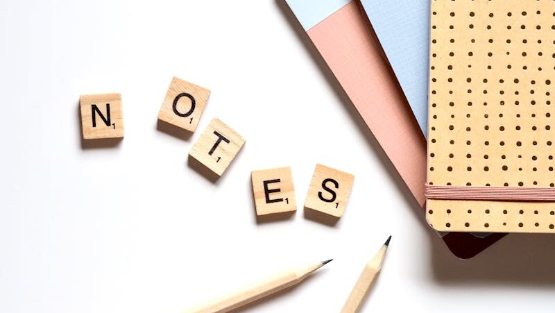 Flat lay of stationery items and Scrabble tiles spelling 'notes' on a white surface.