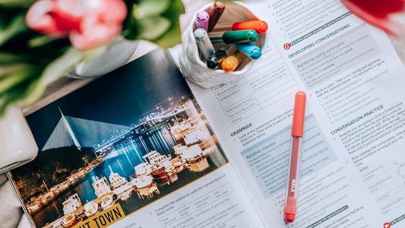 Top view of an open book with pens and markers, surrounded by vibrant flowers, creating a creative workspace vibe.