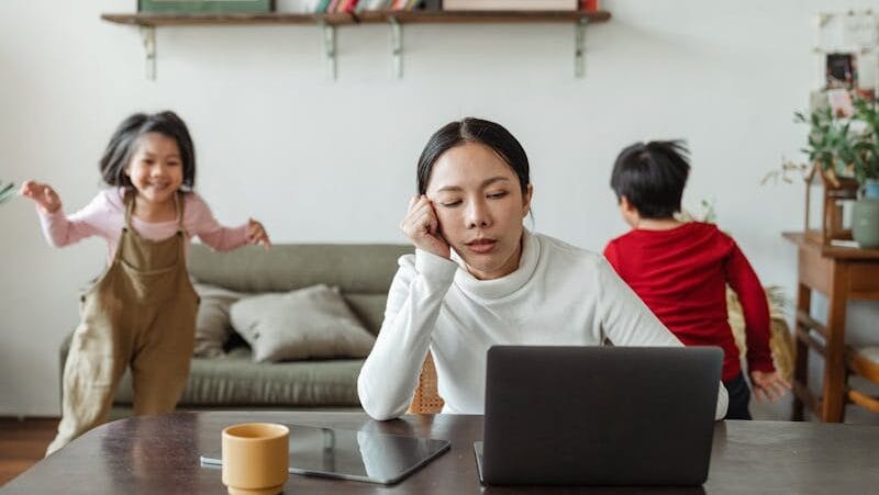 A tired mother working on a laptop while her children play around indoors, highlighting remote work challenges.