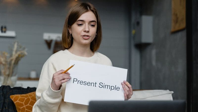 Young woman teaches English grammar online, holding a 'Present Simple' note and pencil.