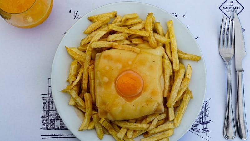 Tasty Portuguese francesinha sandwich topped with egg, surrounded by crispy fries and fresh juice.