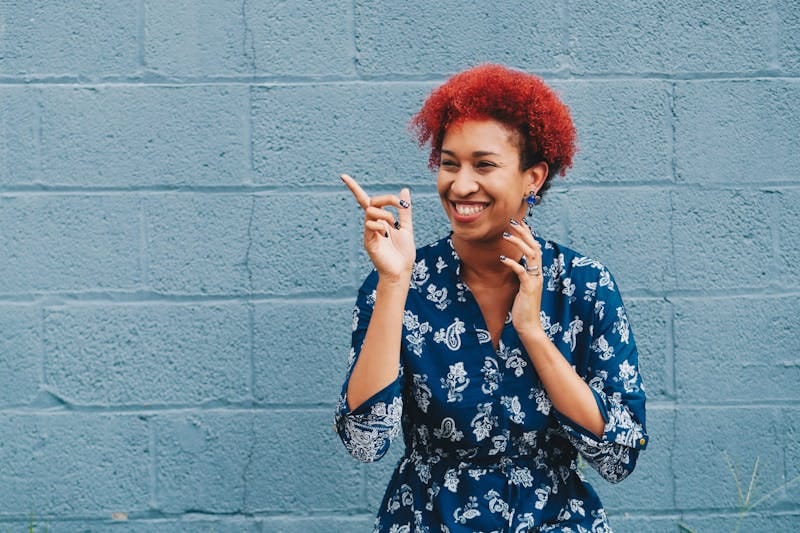 Cheerful lady with red curly hair laughing against a blue brick wall, wearing a patterned dress.
