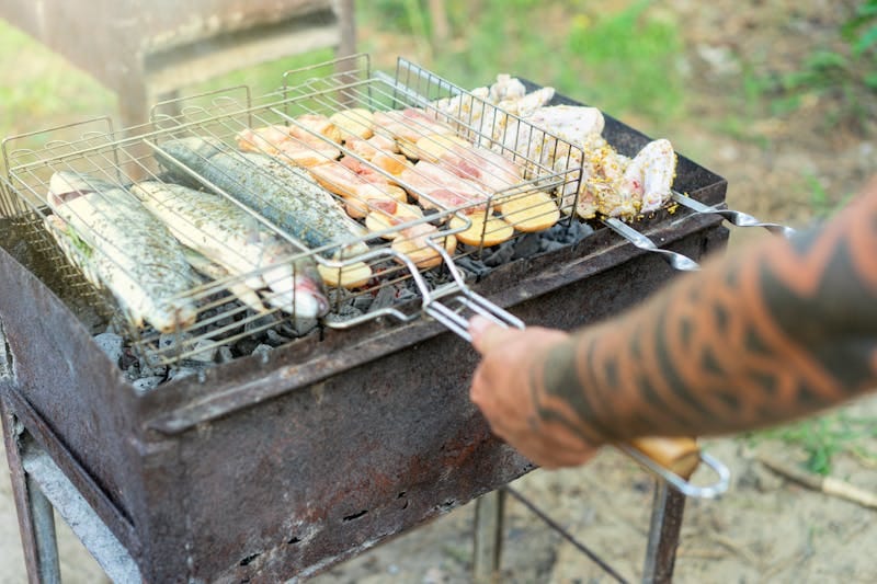 Grilled fish and meats on a barbecue with tattooed arm handling the grill outdoors.