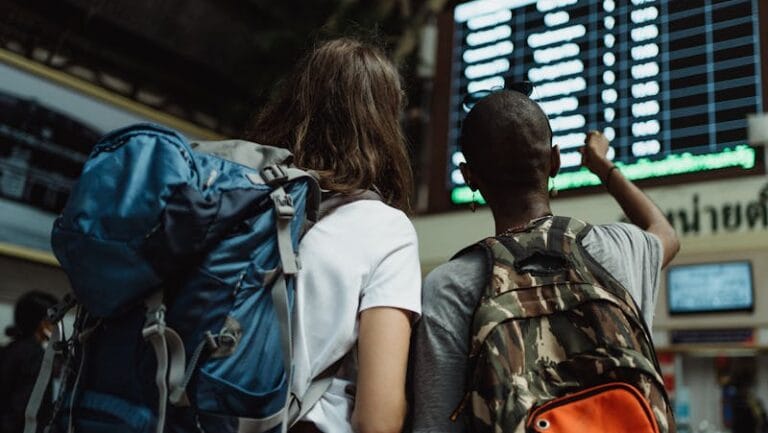 Two backpackers checking the digital board at a train station for travel information.